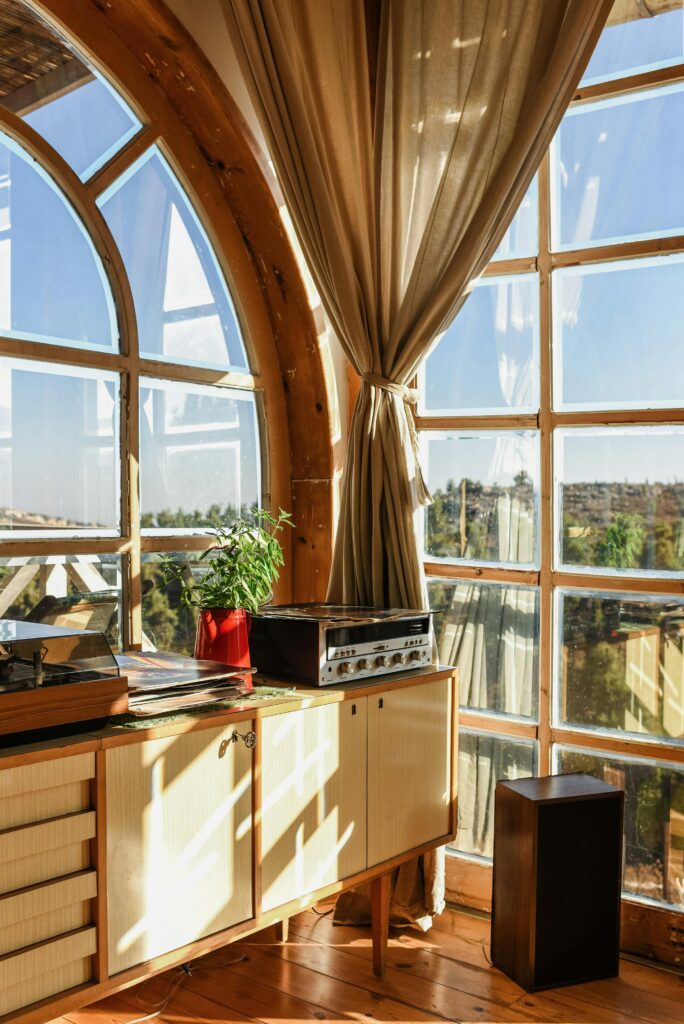 Sunlit cozy corner with a vintage stereo, potted plant, and large arched windows with beige curtains overlooking a peaceful outdoor view.